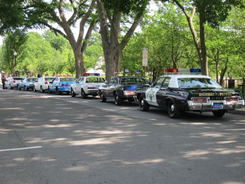 1991 Ford LTD Crown Victoria - long line of classic police cruisers, led by a black-and-white Highway Patrol car, parked curbside under leafy shade trees.