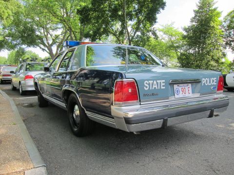 1991 Ford LTD Crown Victoria - Rear view of a two-tone blue and silver State Police sedan with roof light bar parked along a curb beneath shade trees.