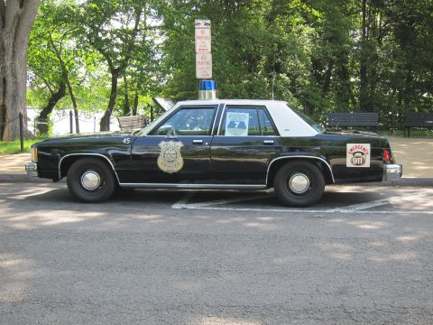 1991 Ford LTD Crown Victoria - Profile of a black police sedan with white roof, gold door shield and Emergency 911 lettering, parked beside a wooded park.