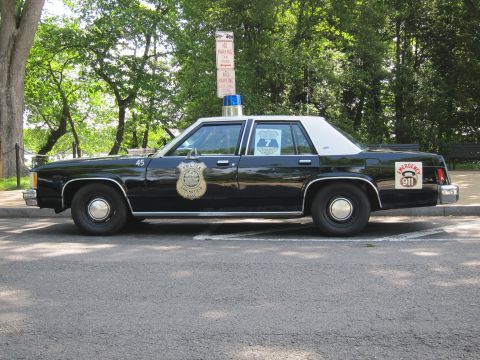 1991 Ford LTD Crown Victoria - black-and-white vintage Ford police sedan parked under trees, marked unit 45 with a gold door shield and a 911 emblem on the trunk.