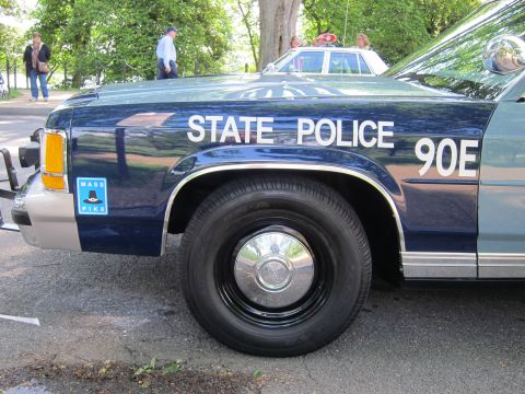 1991 Ford LTD Crown Victoria - Close-up of a dark blue and silver State Police sedan's front fender lettered STATE POLICE 90E with a Mass Pike sticker.
