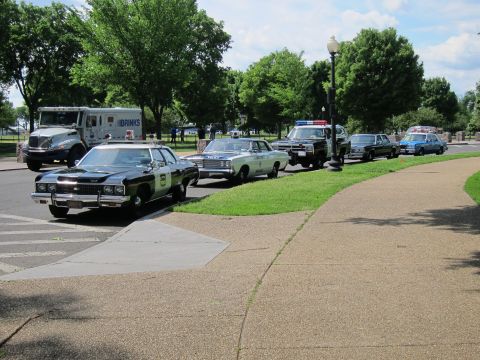 1991 Ford LTD Crown Victoria - row of vintage police cruisers with lightbars and a Brinks armored truck staged along a curving park roadway.