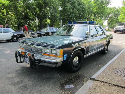 1991 Ford LTD Crown Victoria - dark green and black Ford State Police sedan with front push bar and blue lightbar parked among other classic cruisers.