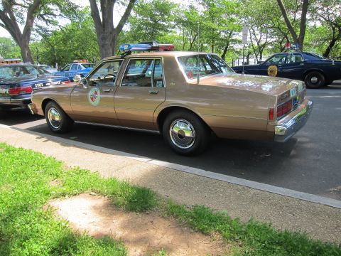 1991 Ford LTD Crown Victoria - tan State Police Chevrolet sedan seen from the rear three-quarter, fitted with a roof lightbar, parked beside other cruisers.