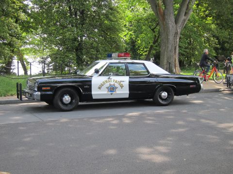 1991 Ford LTD Crown Victoria - black-and-white Highway Patrol Dodge sedan with a single red-and-blue roof light parked on a park road as a cyclist passes.