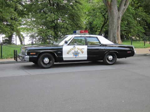 1991 Ford LTD Crown Victoria - Side profile of a black-and-white Dodge Highway Patrol sedan with a gold star door shield and roof beacon, parked by trees.