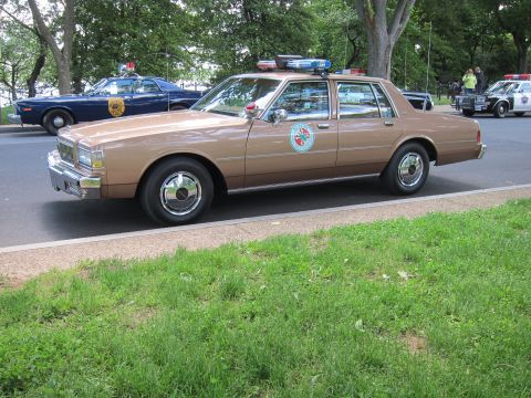 1991 Ford LTD Crown Victoria - tan Chevrolet police sedan with a circular door seal and roof lightbar parked on a park drive among other cruisers.