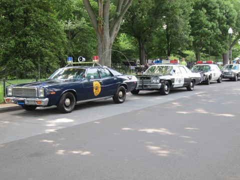 1991 Ford LTD Crown Victoria - line of classic police cruisers along a park road, led by a dark Plymouth sedan with a gold door shield and roof beacons.