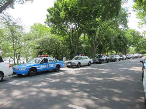 1991 Ford LTD Crown Victoria - long line of vintage police cruisers parked along a tree-shaded park drive, led by a blue-and-white NYPD Chevrolet Caprice.