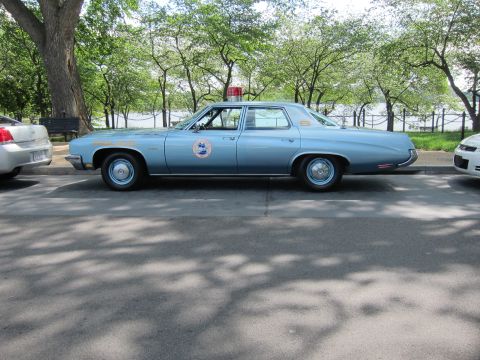 1991 Ford LTD Crown Victoria - Side profile of a light-blue full-size police sedan with a single red roof beacon and door emblem parked along a waterside park drive.