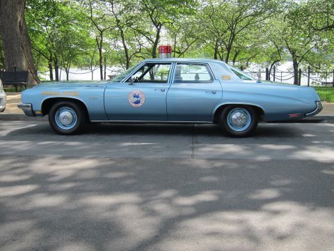 1991 Ford LTD Crown Victoria - Side profile of a light-blue Police-lettered sedan with a circular door seal and red roof beacon parked under park trees.