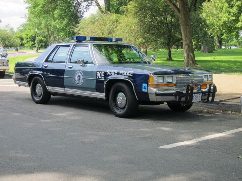 1991 Ford LTD Crown Victoria - dark blue and silver Ford State Police sedan marked 90E with a front push bar and blue roof lights parked on a park road.