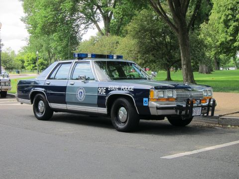 1991 Ford LTD Crown Victoria - blue and silver Ford State Police sedan marked 90E with a chrome push bar and blue roof lights parked under park trees.