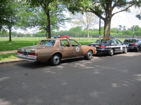 1991 Ford LTD Crown Victoria - tan Chevrolet police sedan with a door seal and roof beacons seen from the rear, parked beside a darker State Trooper cruiser.