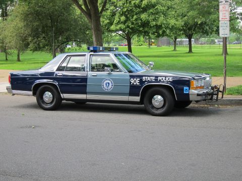 1991 Ford LTD Crown Victoria - dark blue and silver Ford State Police sedan marked 90E with a front push bar and blue roof lights parked near a no-stopping sign.