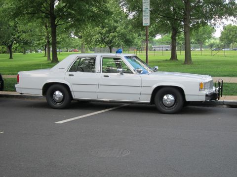 1991 Ford LTD Crown Victoria - white full-size four-door sedan outfitted as a police car with a blue roof beacon, parked on a curb beside a grassy park.
