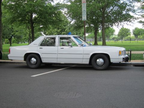 1991 Ford LTD Crown Victoria - white full-size four-door police sedan with a single blue roof light and push bar, parked along a curb near a green park.