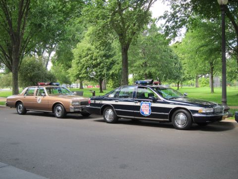1991 Ford LTD Crown Victoria - tan sedan parked nose-to-tail with a black-and-white 'State Trooper' sedan bearing a shield emblem on a tree-lined park road.