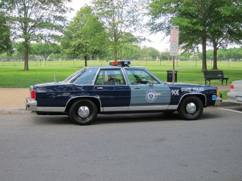 1991 Ford LTD Crown Victoria - two-tone navy-and-silver State Police sedan marked '90E STATE POLICE' with roof lights, parked on a curb beside a park.