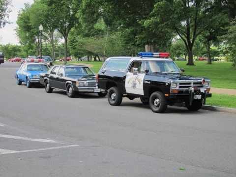 1991 Ford LTD Crown Victoria - black-and-white highway patrol SUV with light bar and brush guard leads a line of vintage police cars on a park road.