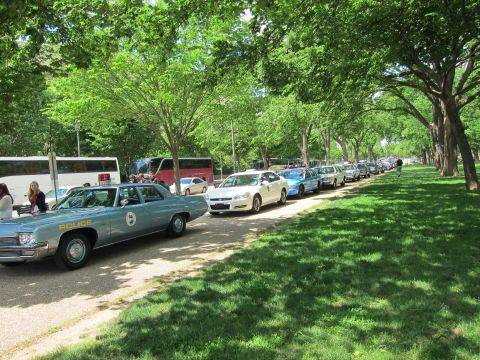 1991 Ford LTD Crown Victoria - light-blue vintage police sedan marked 'POLICE' heads a long line of patrol cars parked along a shaded tree-lined path.