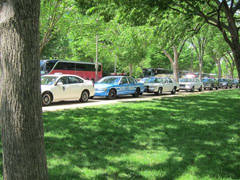1991 Ford LTD Crown Victoria - blue-and-white NYPD police sedan heads a line of parked patrol cars along a path bordered by a grassy lawn and trees.