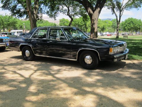 1991 Ford LTD Crown Victoria - dark navy full-size four-door sedan with chrome trim and a roof spotlight, parked on gravel beneath shade trees in a park.