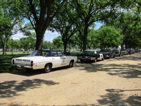 1991 Ford LTD Crown Victoria - white 'State Police' sedan with rear light bar leads a line of vintage patrol cars parked on a shaded gravel park lane.