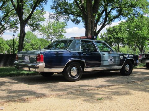 1991 Ford LTD Crown Victoria - Rear-quarter view of a navy-and-silver 'State Police' sedan marked '90E' with seal emblem and roof light bar, parked under trees.