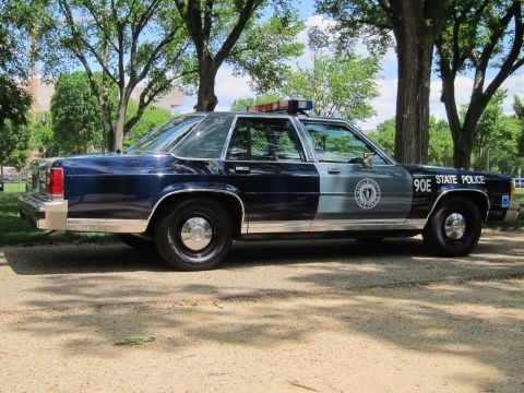 1991 Ford LTD Crown Victoria - Side profile of a navy-and-silver 'State Police' sedan marked '90E' with roof lights and seal emblem, parked on gravel.