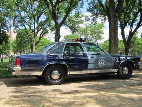 1991 Ford LTD Crown Victoria - navy-and-silver 'State Police' sedan marked '90E' with circular seal and roof light bar, parked on gravel beneath trees.