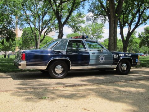 1991 Ford LTD Crown Victoria - Front-quarter view of a navy-and-silver 'State Police' sedan marked '90E' with seal emblem and roof lights, parked under trees.