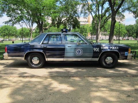 1991 Ford LTD Crown Victoria - Full side view of a navy-and-silver 'State Police' sedan marked '90E STATE POLICE' with seal emblem, parked on gravel in a park.