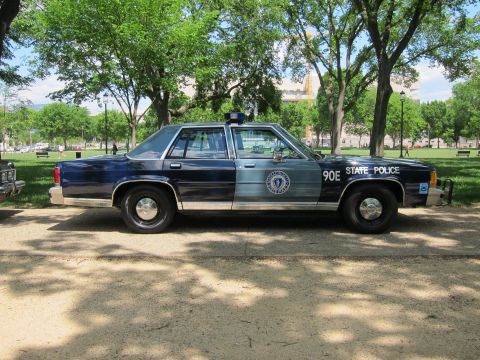 1991 Ford LTD Crown Victoria - Side profile of a navy-and-silver 'State Police' sedan marked '90E' with circular seal and roof light, parked under shade trees.