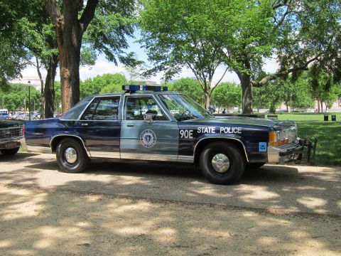 1991 Ford LTD Crown Victoria - navy-and-silver 'State Police' sedan marked '90E' with twin blue roof lights and seal emblem, parked on gravel in a park.