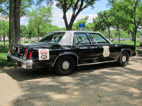 1991 Ford LTD Crown Victoria - Rear-quarter view of a black full-size police sedan with an 'Emergency 911' decal, badge emblem and blue roof light, parked in a park.