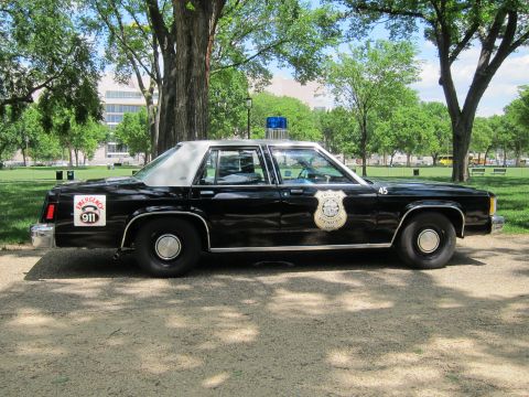 1991 Ford LTD Crown Victoria - Side view of a black full-size police sedan bearing an 'Emergency 911' decal, badge emblem and blue roof light, parked on gravel.