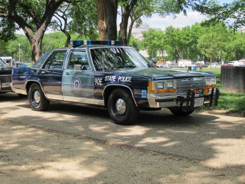1991 Ford LTD Crown Victoria - dark green and gray Ford LTD Crown Victoria marked '90E State Police' sits parked on a dirt lot among shade trees.