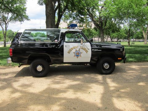 1991 Ford LTD Crown Victoria - Profile of a black and white 'Highway Patrol' Dodge Ramcharger SUV with a seven-point star door emblem, parked in a park.