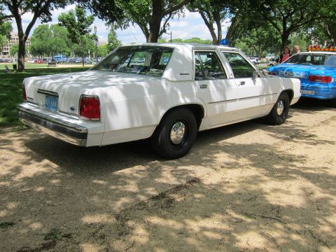 1991 Ford LTD Crown Victoria - Rear three-quarter view of a white Ford LTD Crown Victoria police sedan parked beside a blue cruiser in a shaded park.