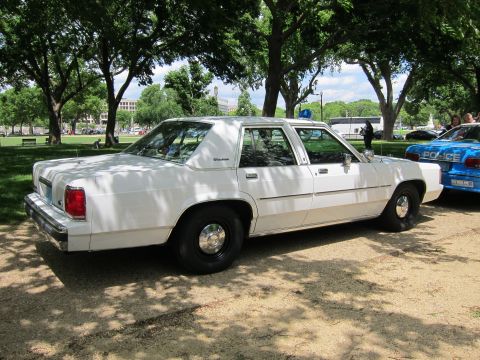 1991 Ford LTD Crown Victoria - white Ford LTD Crown Victoria sedan parked on a dirt lot beside a blue 'Police' car under shade trees.