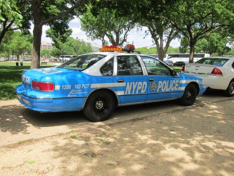 1991 Ford LTD Crown Victoria - Rear three-quarter view of a blue and white Chevrolet Caprice marked 'NYPD Police 103 PCT' with roof lights, parked under trees.