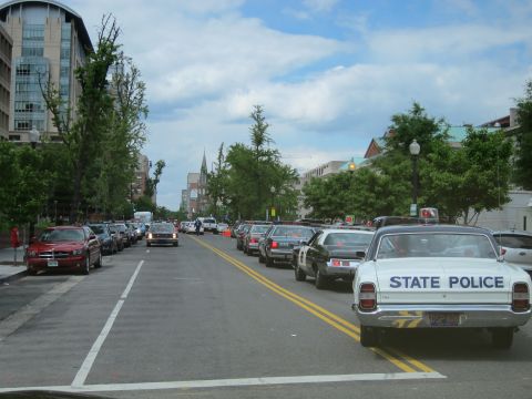 1991 Ford LTD Crown Victoria - Rear view of a white vintage 'State Police' sedan driving down a tree-lined two-way city street past parked cars.