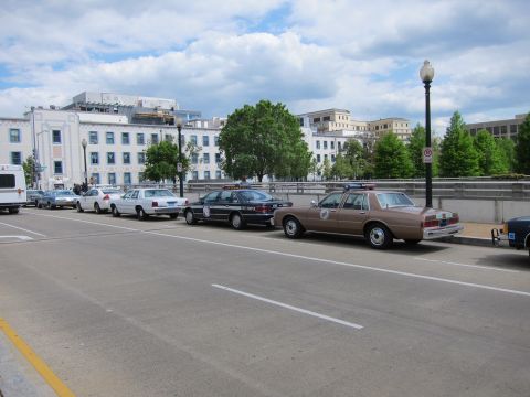 1991 Ford LTD Crown Victoria - row of police cruisers, including a brown Chevrolet Caprice, parked along a city street beside multi-story buildings.