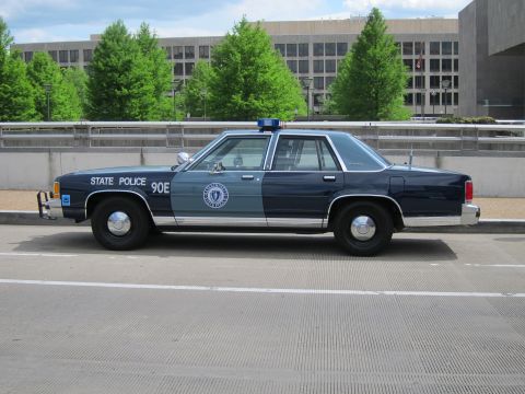 1991 Ford LTD Crown Victoria - Profile of a green and gray Ford LTD Crown Victoria marked '90E State Police' parked on pavement before a concrete building.