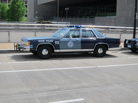 1991 Ford LTD Crown Victoria - Front three-quarter view of a green and gray '90E State Police' Ford sedan with push bar, parked on a city street.