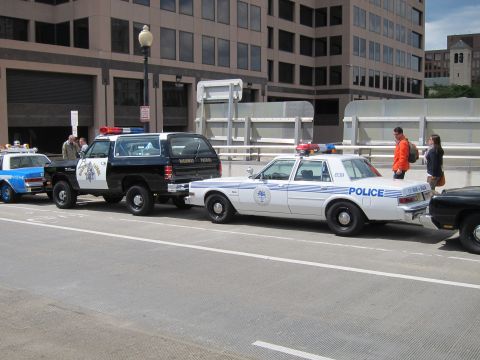 1991 Ford LTD Crown Victoria - black 'Highway Patrol' SUV and a white Dodge 'Police' sedan parked along a city street with pedestrians and buildings.