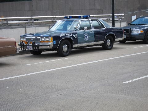 1991 Ford LTD Crown Victoria - Front three-quarter view of a green and gray '90E State Police' Ford sedan with push bar, parked on a downtown street.
