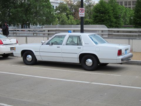 1991 Ford LTD Crown Victoria - white Ford LTD Crown Victoria police sedan with a single blue beacon parked along a city street near a no-parking sign.