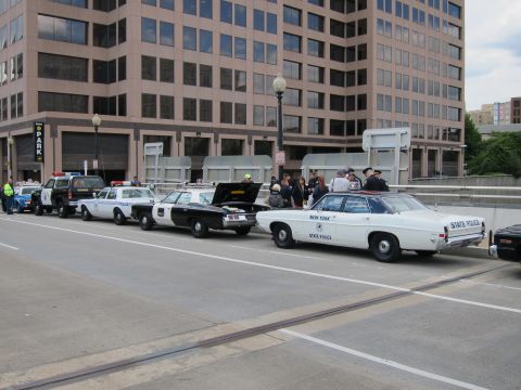 1991 Ford LTD Crown Victoria - line of vintage police cars, including a white 'New York State Police' sedan, parked along a street by a glass office tower.